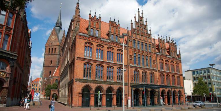 Backsteingebäude mit neugotischer Fassade in Hannover, im Hintergrund die Marktkirche mit spitzem Turm.