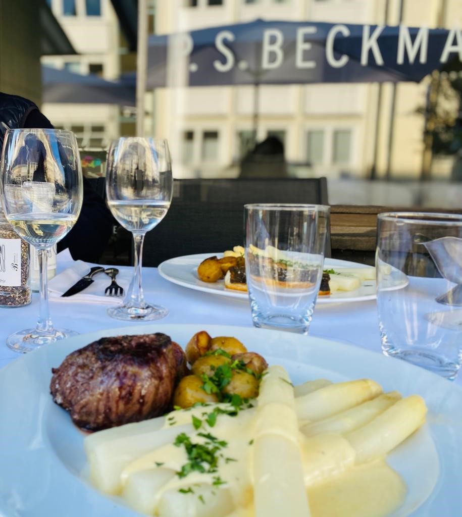 Beckmanns Weinhaus, Tisch mit weißer Tischdecke vor Fenster mit sonniger Spiegelung, zwei Weingläser auf dem Tisch. ein Teller mit Steak und Spargel im Vordergrund.