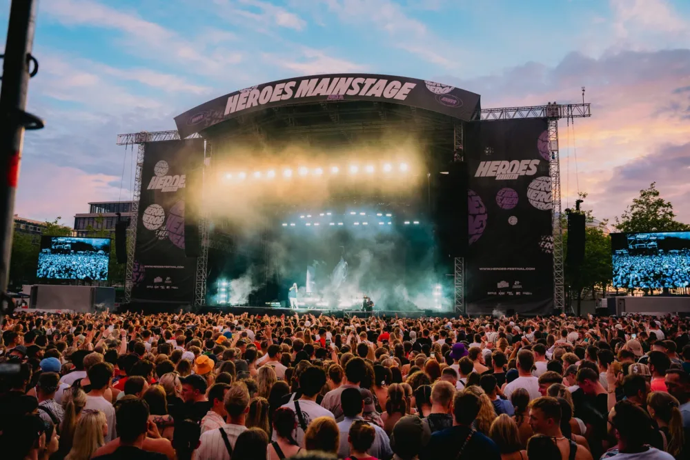 A large crowd celebrates in front of the illuminated "Heroes Mainstage" at sunset while an artist performs.