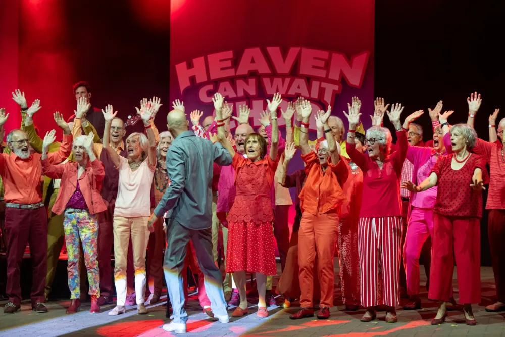 一个由身穿红色服装的老人组成的合唱团在舞台上高歌一曲，并举起手臂，上面写着 "天堂可以等待"。