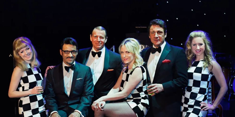 A group of elegantly dressed people pose on a stage. The women are wearing black and white chequerboard dresses with white boots.