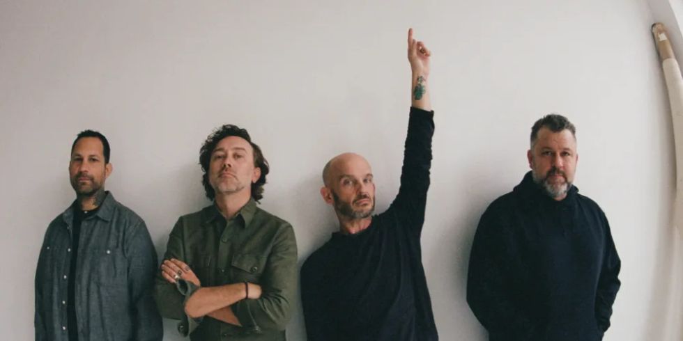 Four men stand in front of a white wall. Three look serious, one raises a hand upwards. They are wearing dark, casual clothes.
