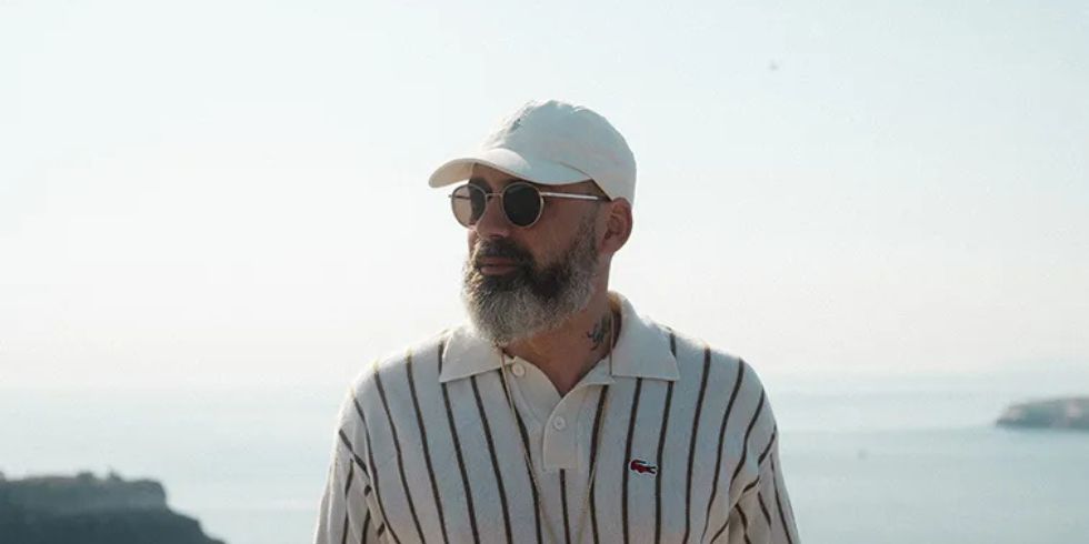 Ein Mann mit Bart, Sonnenbrille und weißer Kappe steht entspannt vor einer Meereskulisse mit Klippen und blauem Himmel.