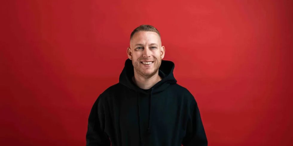 A smiling man in a black hoodie stands in front of a red background and puts his hands in his pockets.