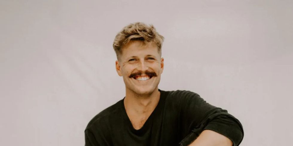 Man with a moustache sits smiling on a stool in front of a light-coloured background, wearing a black outfit.
