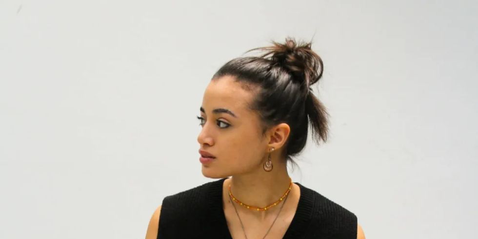 梳着发髻的年轻女子坐在凳子上，身穿黑色毛衣和宽大的长裤，目光投向一旁。