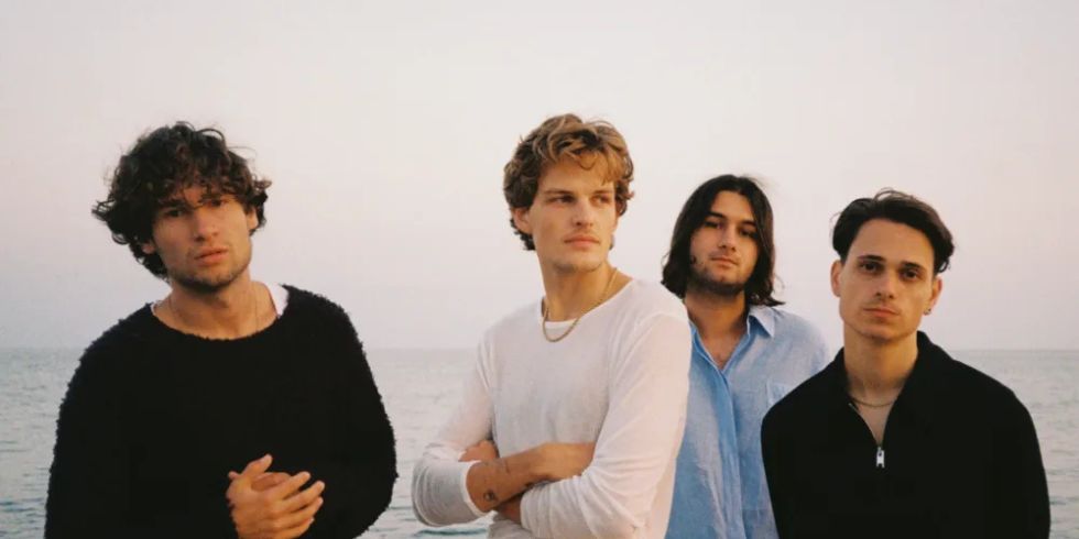 Vier junge Männer stehen am Strand vor dem Meer, in stilvoller Kleidung und Abendlicht.