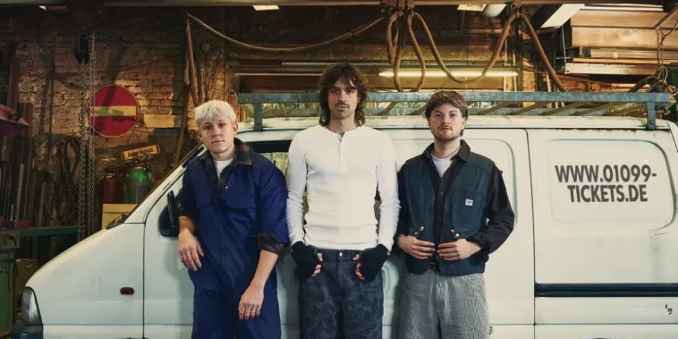 Drei junge Männer stehen vor einem weißen Transporter mit der Webadresse „www.01099-tickets.de“ in einer Werkstatt.