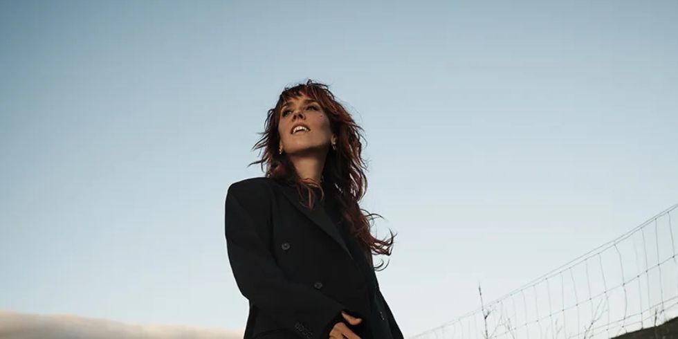 Woman in a black suit stands on a country lane, smiling and looking up at the sky in the evening light.