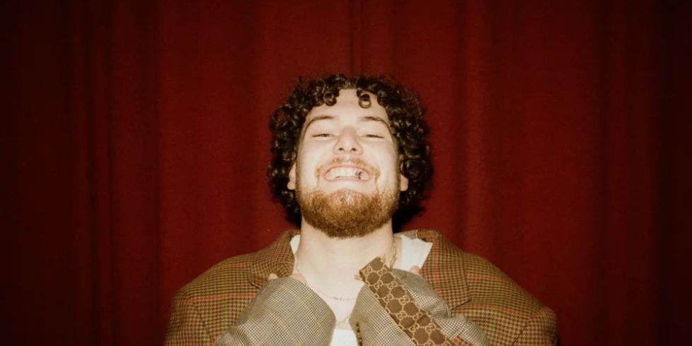 Laughing man with curly hair and beard wearing a chequered oversize blazer in front of a red curtain.