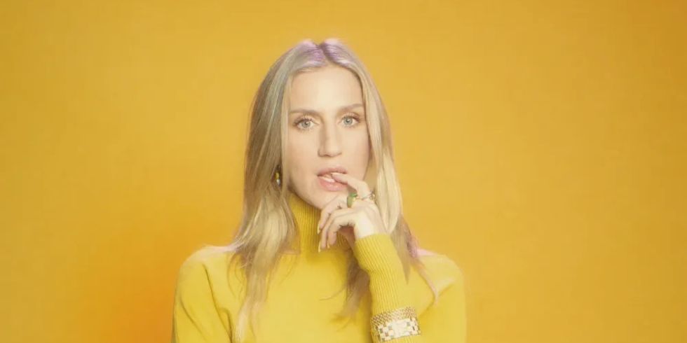 身着黄色连衣裙的女子站在黄色背景前，手放在嘴唇上，若有所思地看着镜头。