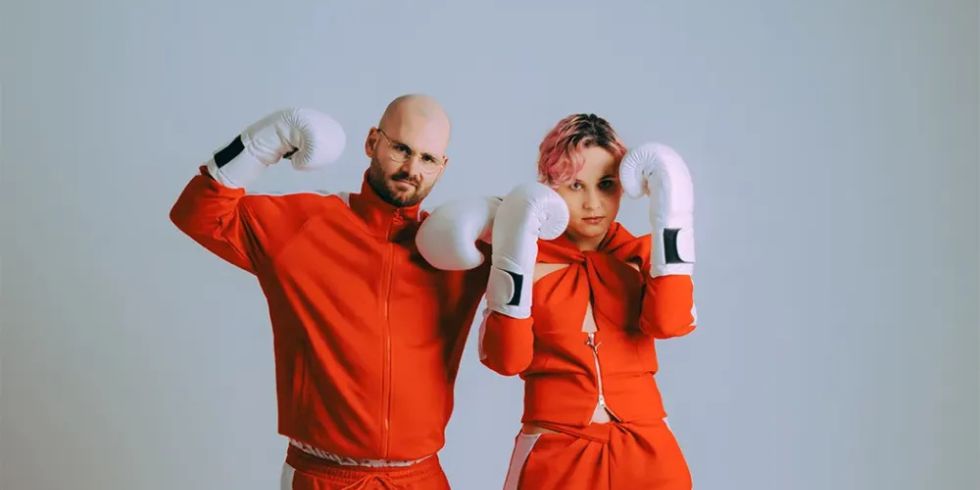 Mann und Frau in roten Trainingsanzügen tragen weiße Boxhandschuhe und posieren kämpferisch vor hellem Hintergrund.