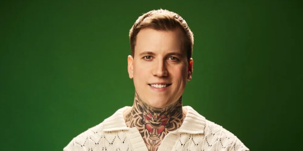 Smiling man with tattooed arms and neck sits in front of a green background in a light-coloured knitted shirt.