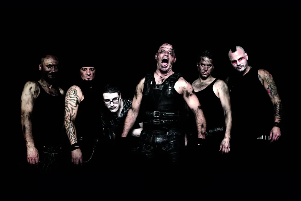 Sechs Männer in düsterem Bühnenoutfit mit Schminke und Tattoos posieren kraftvoll vor schwarzem Hintergrund.