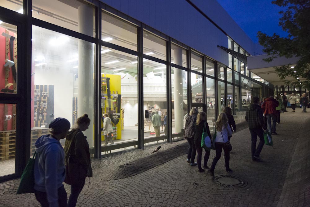 People walk along a glazed façade in the evening with a view of an illuminated art exhibition.