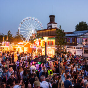 Großes Volksfest am Abend mit Menschenmenge, beleuchteten Buden, Riesenrad und Feststimmung bei Sommerdämmerung.