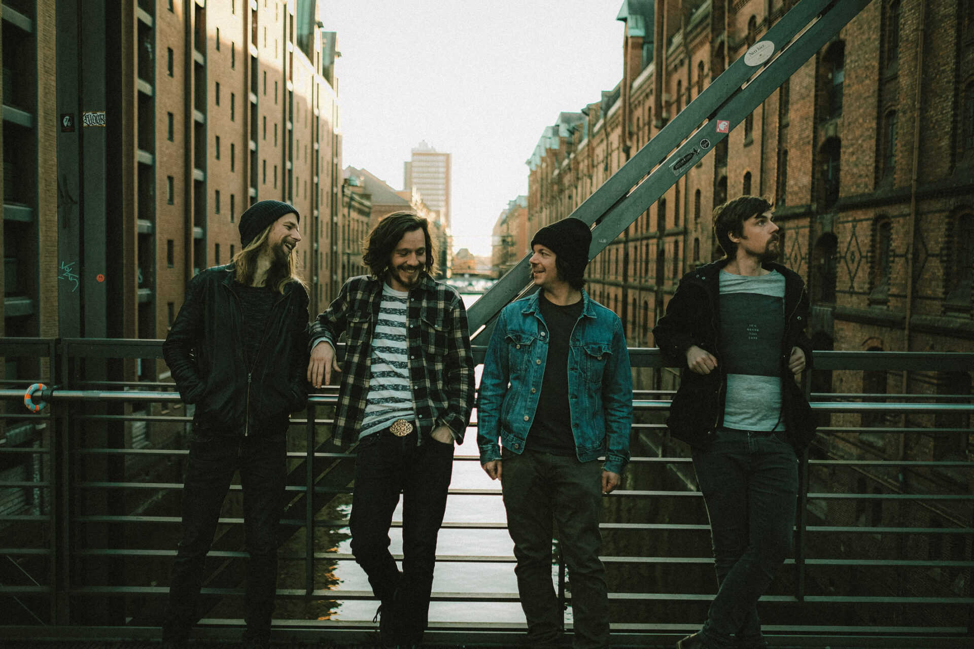 Four-piece band stands laughing on a bridge between brick buildings in an urban environment in daylight.