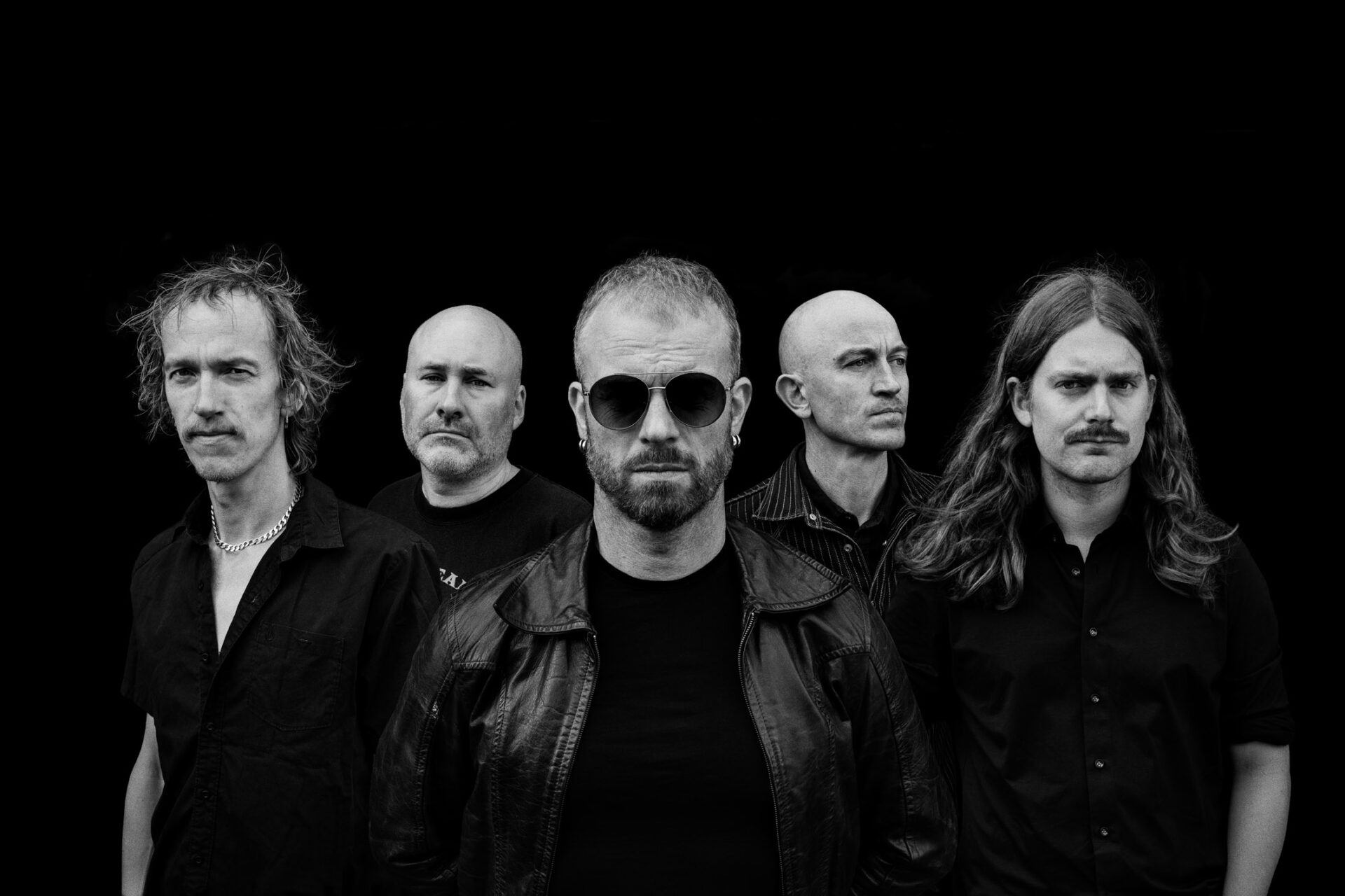 Five-piece rock band in black and white, looking serious in front of a black background, central figure with sunglasses.