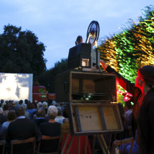 Open-Air-Kino mit Filmvorführung auf Leinwand, Zuschauer sitzen auf Stühlen im Park bei Abenddämmerung.