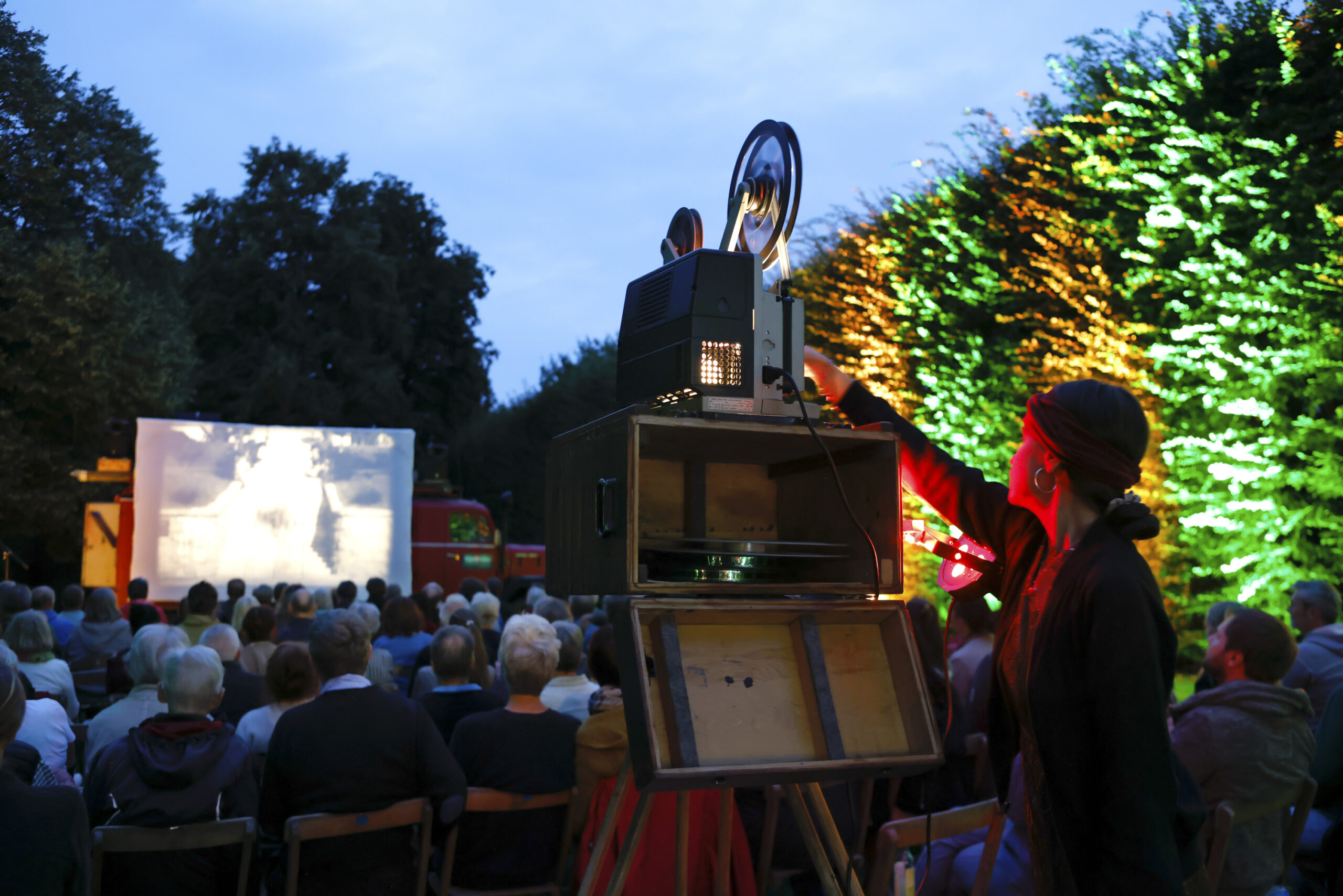 Open-air cinema with film screening on a screen, audience sitting on chairs in the park at dusk.