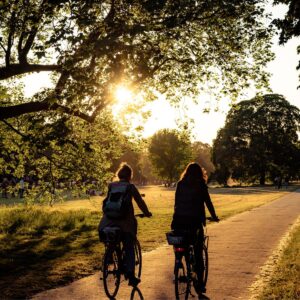 Zwei Radfahrerinnen bei Sonnenuntergang auf einem Parkweg, umgeben von Bäumen und warmem Licht.