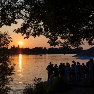 Menschen sitzen am Ufer bei Sonnenuntergang, genießen die Aussicht auf den See mit goldener Spiegelung.