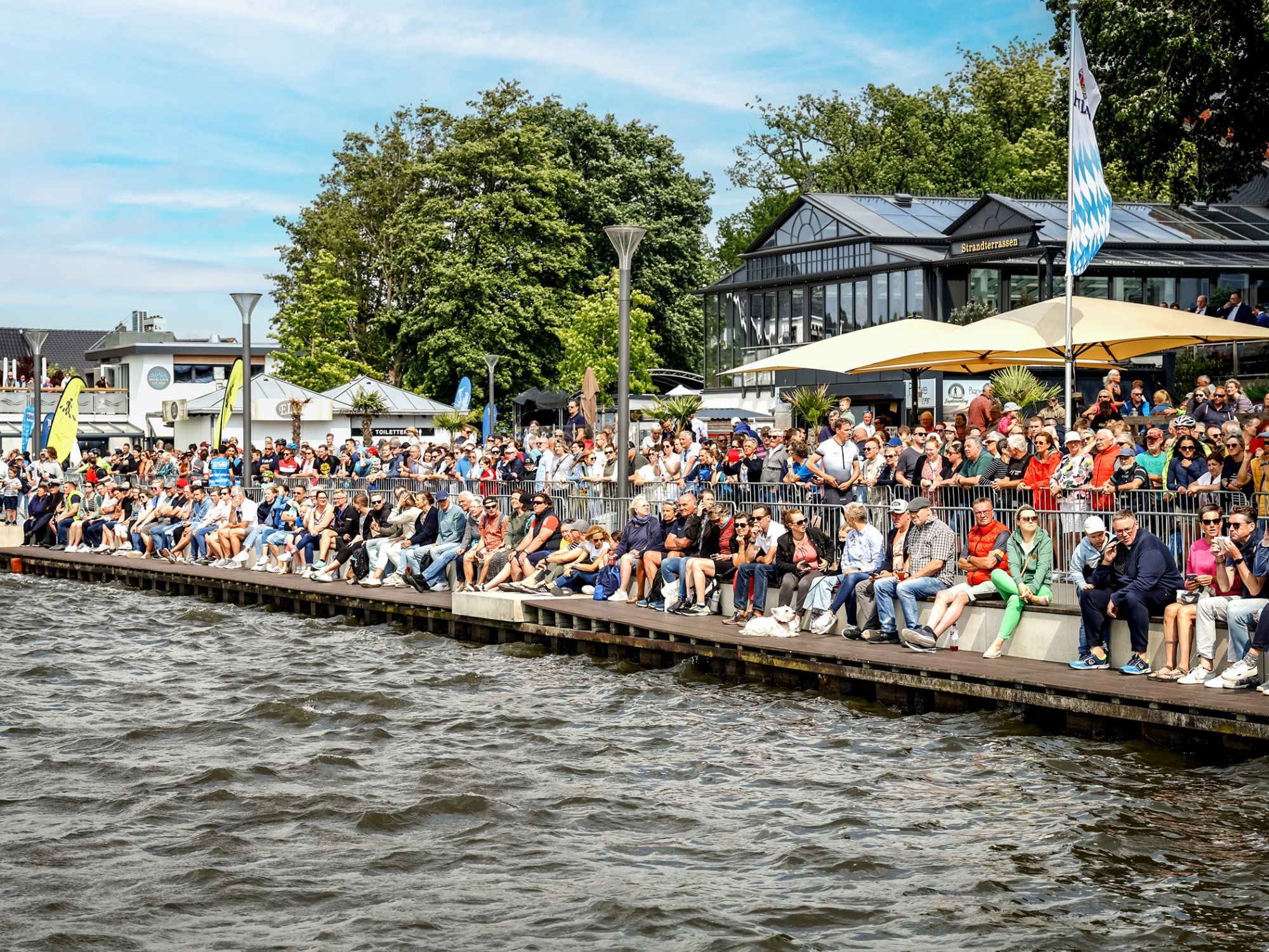 Spectators sit tightly packed on the promenade and watch a sporting event on the water.