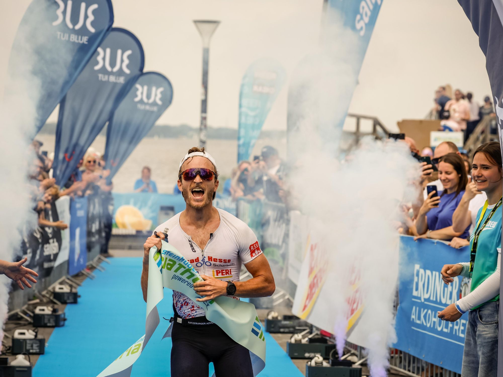 Triathlete cheers at the finish line on a blue carpet, surrounded by smoke and cheering spectators.