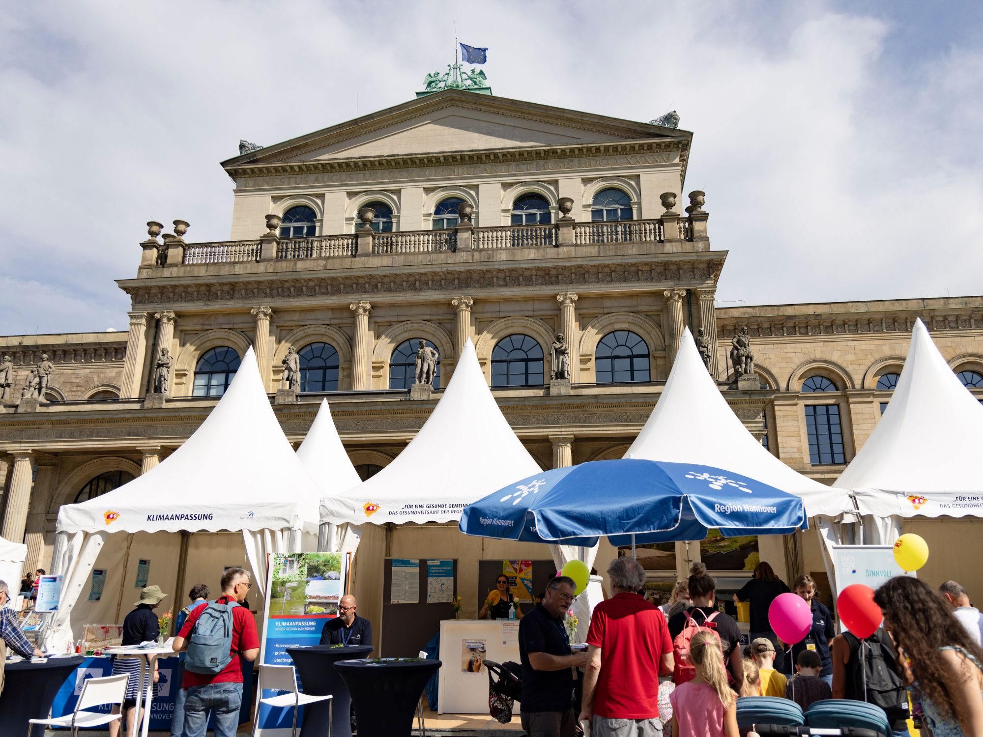 Visitors in front of white information stands and pavilions on Hanover's Opernplatz in sunny weather.