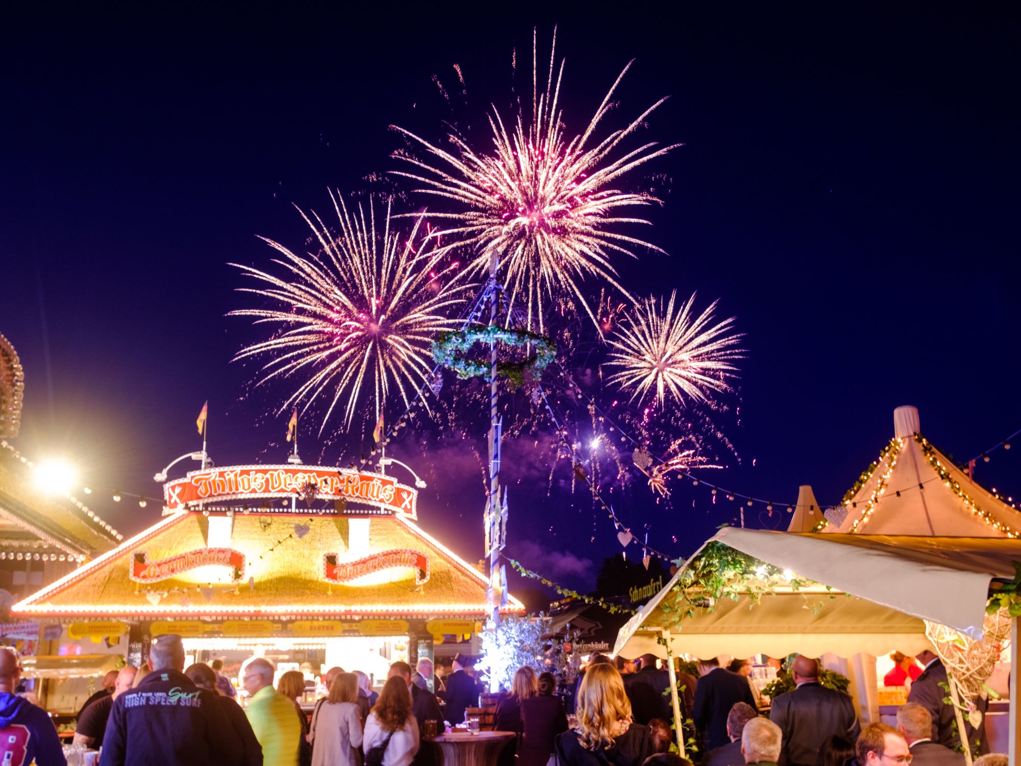 Fireworks over a festively illuminated folk festival with crowds of people and decorated stalls at night.