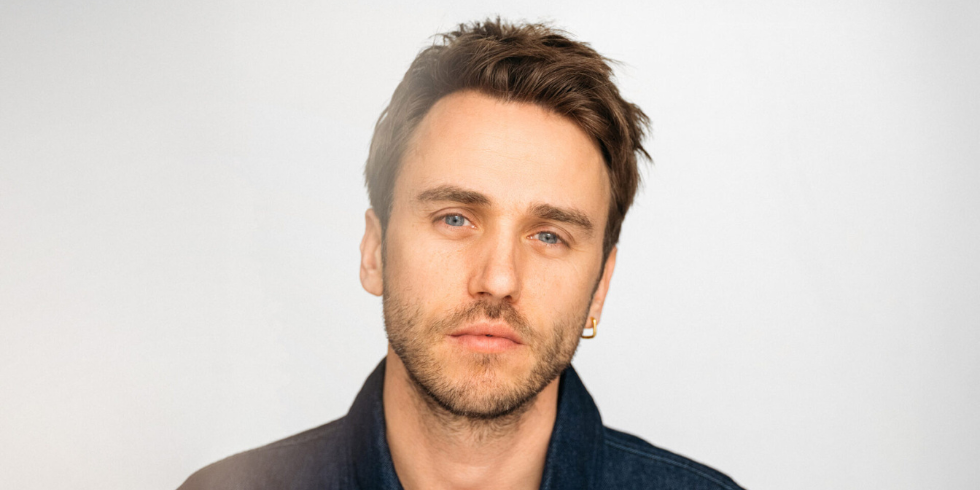 Portrait of a man with a serious look, beard and earring against a light background in a dark denim jacket.