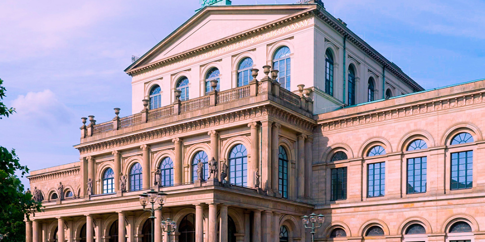 Die Staatsoper Hannover bei Tageslicht, mit Regenbogenflagge auf dem Dach und Menschen auf dem Vorplatz.
