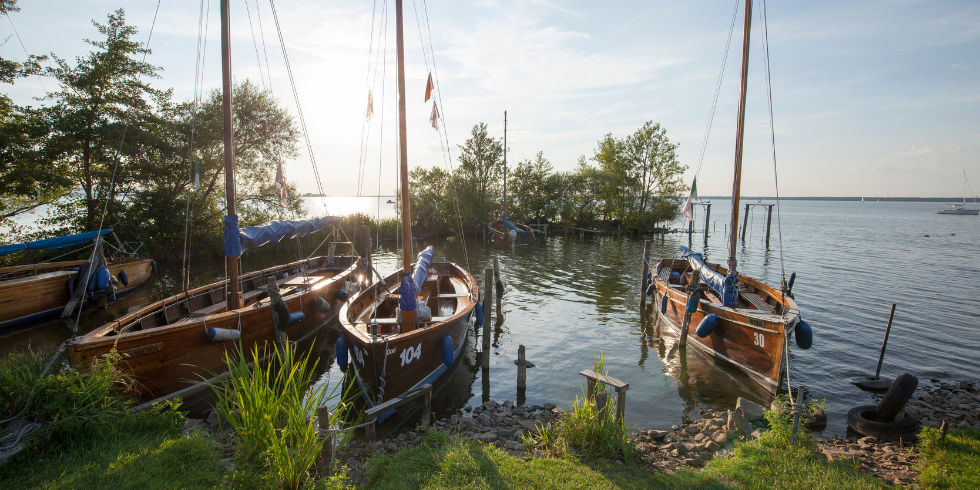 Mehrere Holzsegelboote liegen am Ufer eines ruhigen Sees bei tiefstehender Sonne.