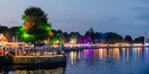 Abendliches Fest am See mit bunten Lichtern, vielen Menschen und stimmungsvoller Spiegelung im Wasser.