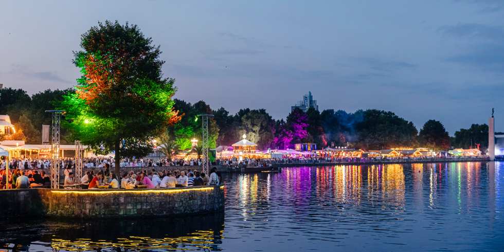 Abendliches Fest am See mit bunten Lichtern, vielen Menschen und stimmungsvoller Spiegelung im Wasser.