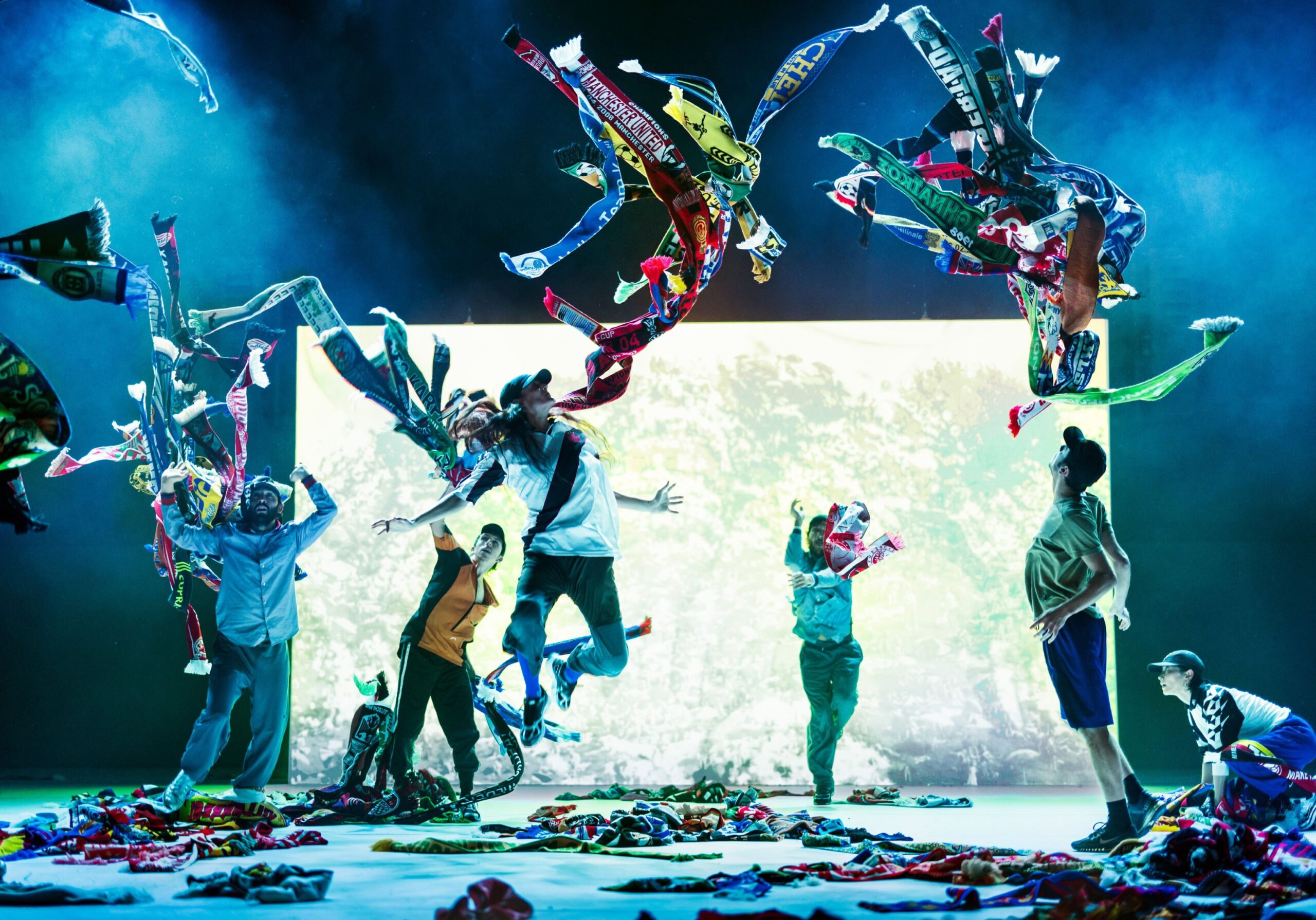 Dancers twirl football scarves through the air, a dynamic scene on a colourfully lit stage.
