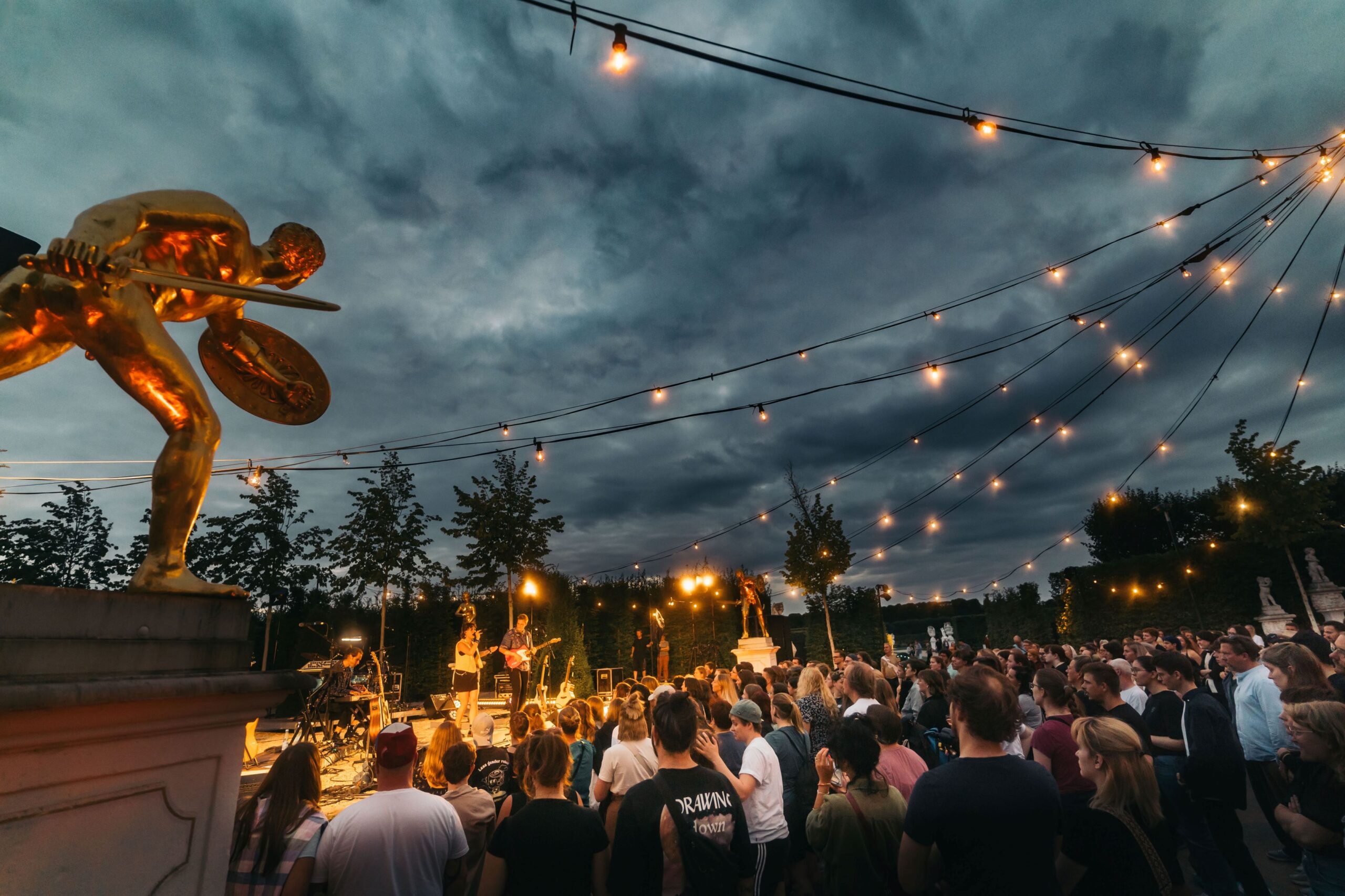 傍晚时分，观众们在花园里欣赏露天音乐会，音乐会上点缀着童话灯光和金色雕像。