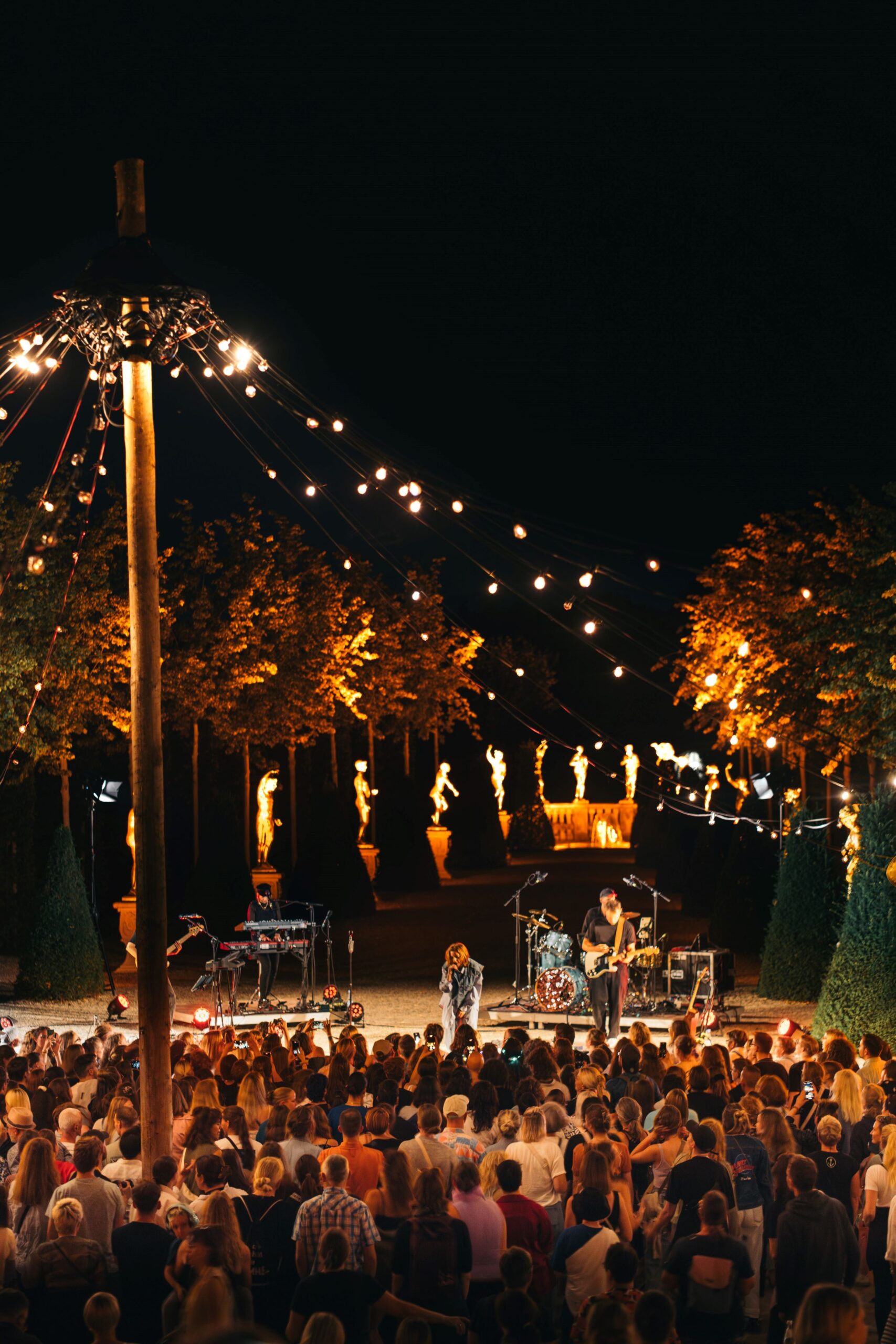 夜间在灯火通明的花园中举行露天音乐会,观众在舞台前与音乐家和童话灯一起欣赏演出。